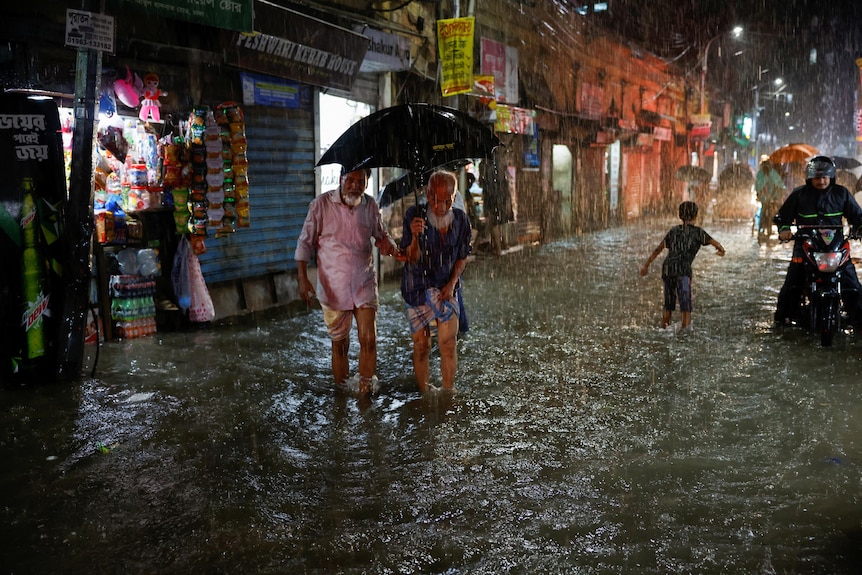 Dos hombres mayores caminan en el agua hasta los tobillos bajo un paraguas mientras otros caminan o andan en bicicleta detrás de ellos.
