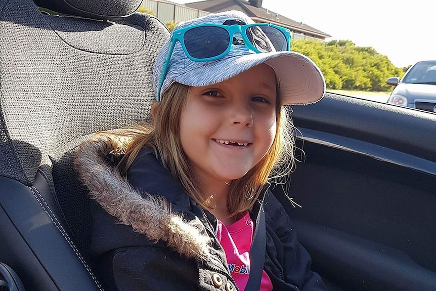 A mid shot of eight-year-old junior drag racer Anita Board sitting in a car wearing a hat smiling for a photo.