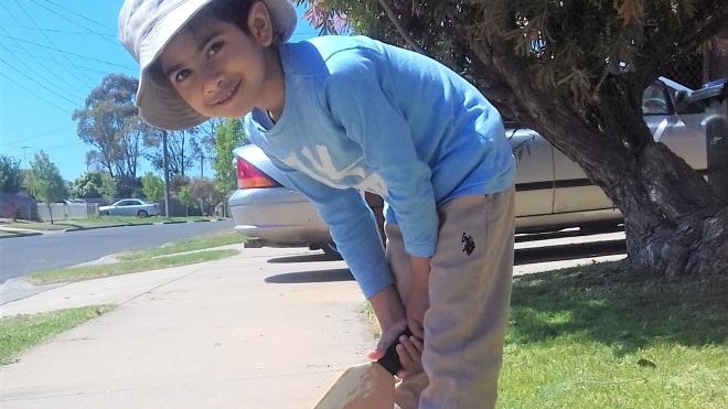 A smiling five-year-old boy stands on a footpath and holds a cricket bat in both hands.