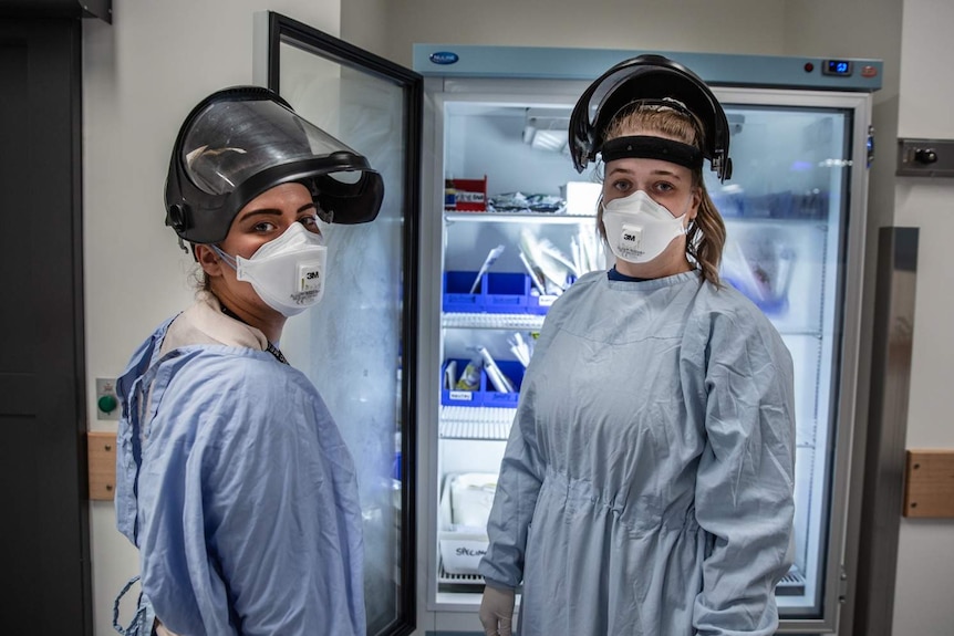 Two women wearing long protective gowns and medical masks look at the camera