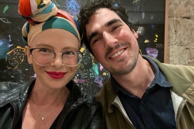 A woman wearing glasses and a headscarf and a man. Both are smiling