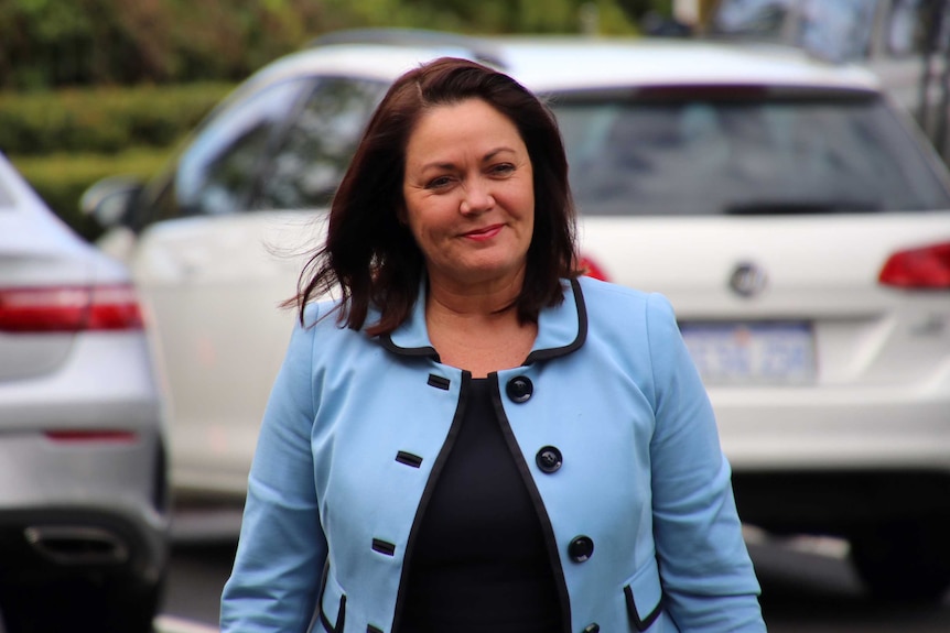 A mid shot of a smiling Liza Harvey walking in front of parked cars wearing a blue jacket.