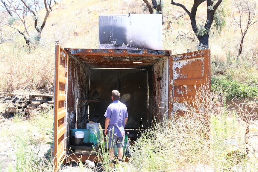 A man faces a burnt out shipping container sitting on a plot of rural land.