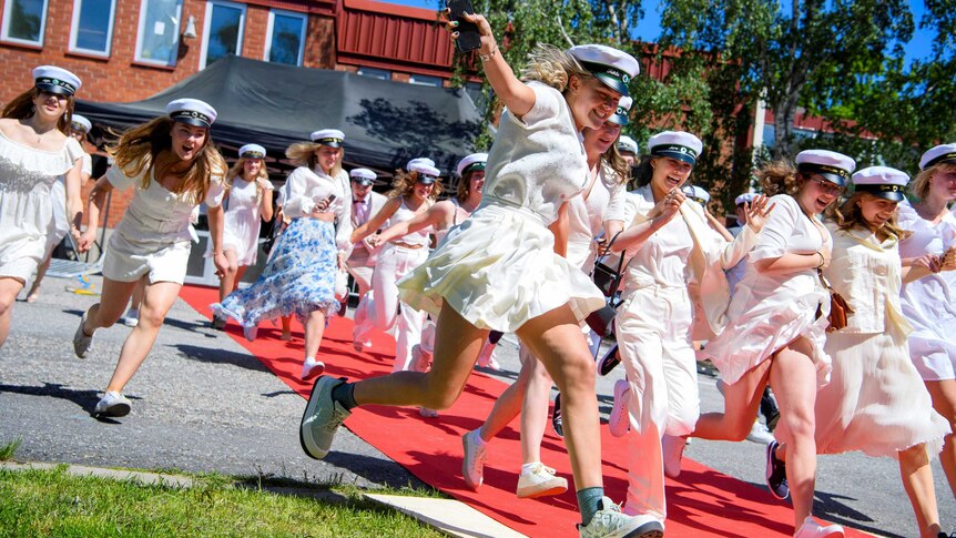 Young women in white dresses and sailor caps running out of a school building