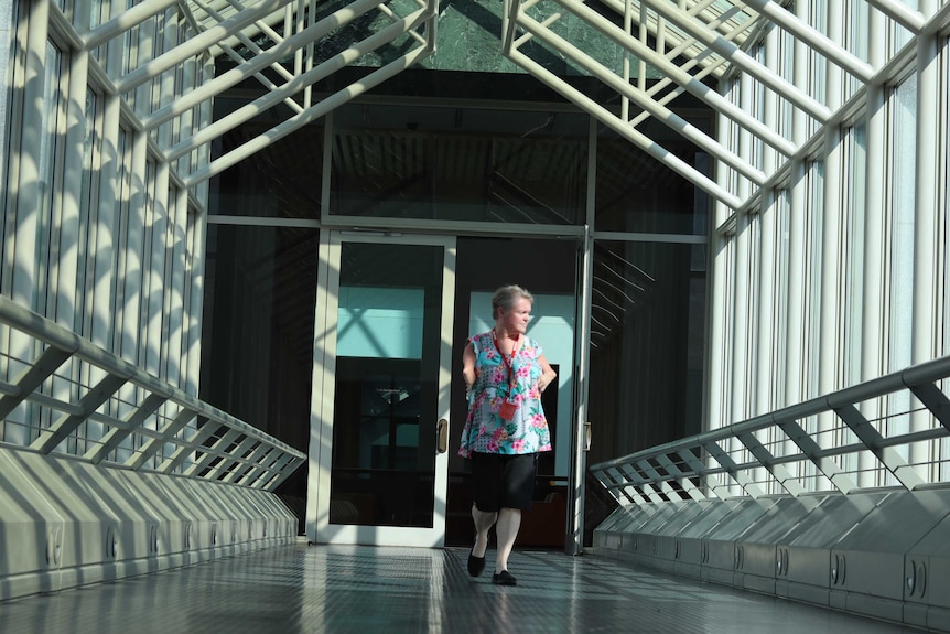 A woman with shortened arms walks through a corridor which sends shadows through the area