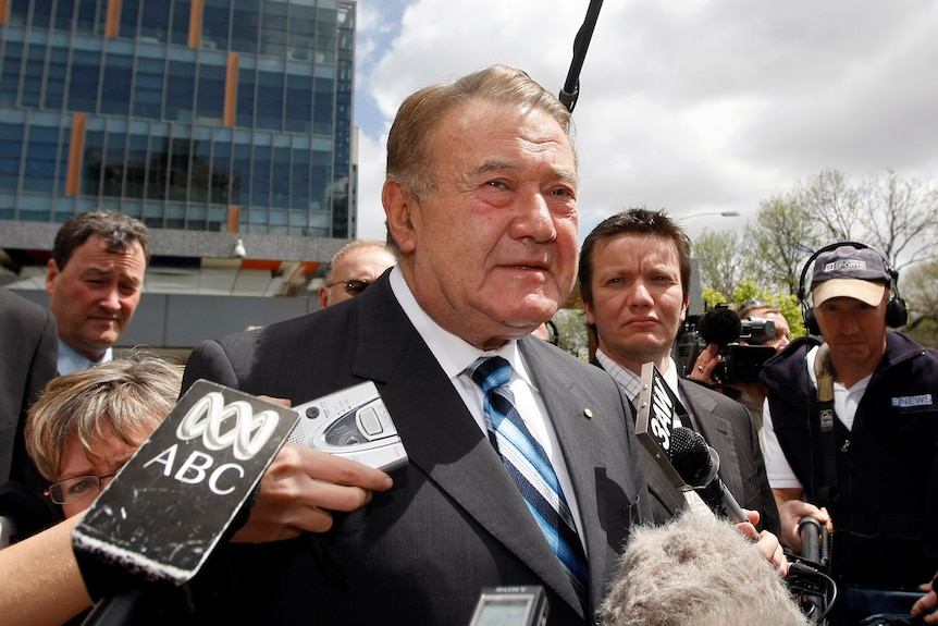 An older man wearing a suit and tie is swarmed by reporters with microphones and tape recorders.