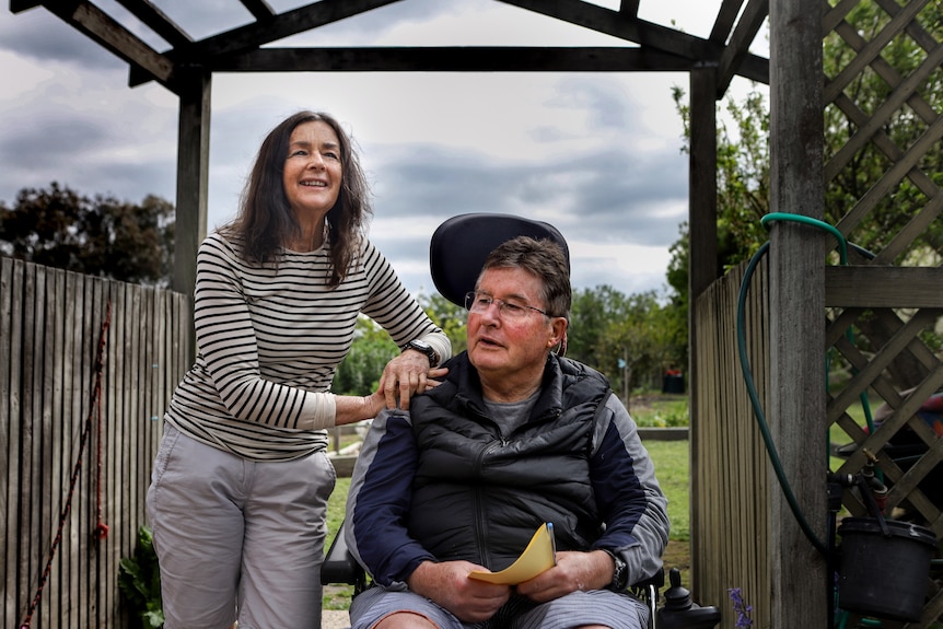 Woman stands next to man in wheelchair under wooden frame with vegetable garden visible in background