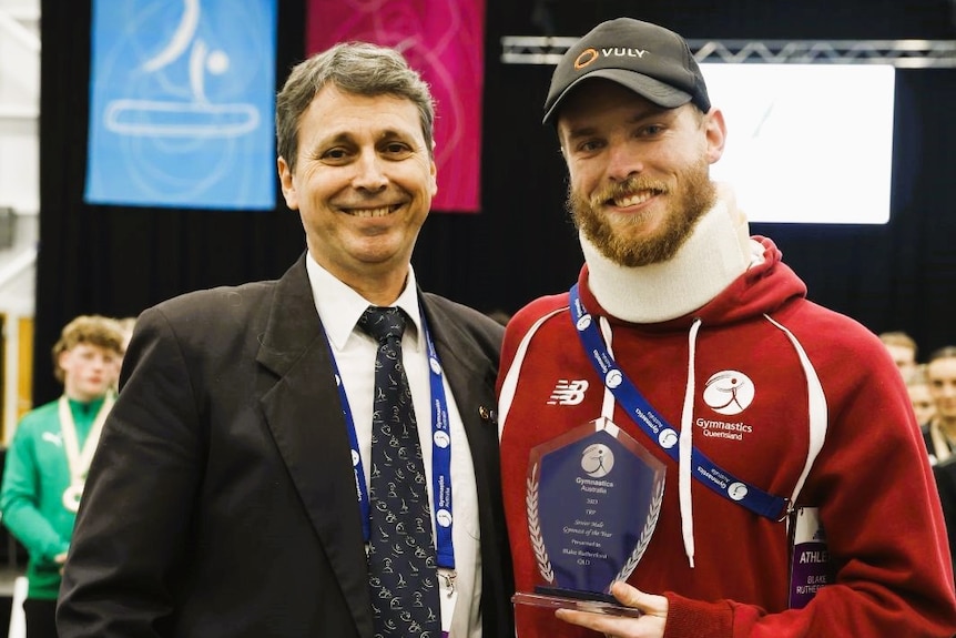 A man in a suit and a man wearing a tracksuit and cap in a neck brace holding an award.