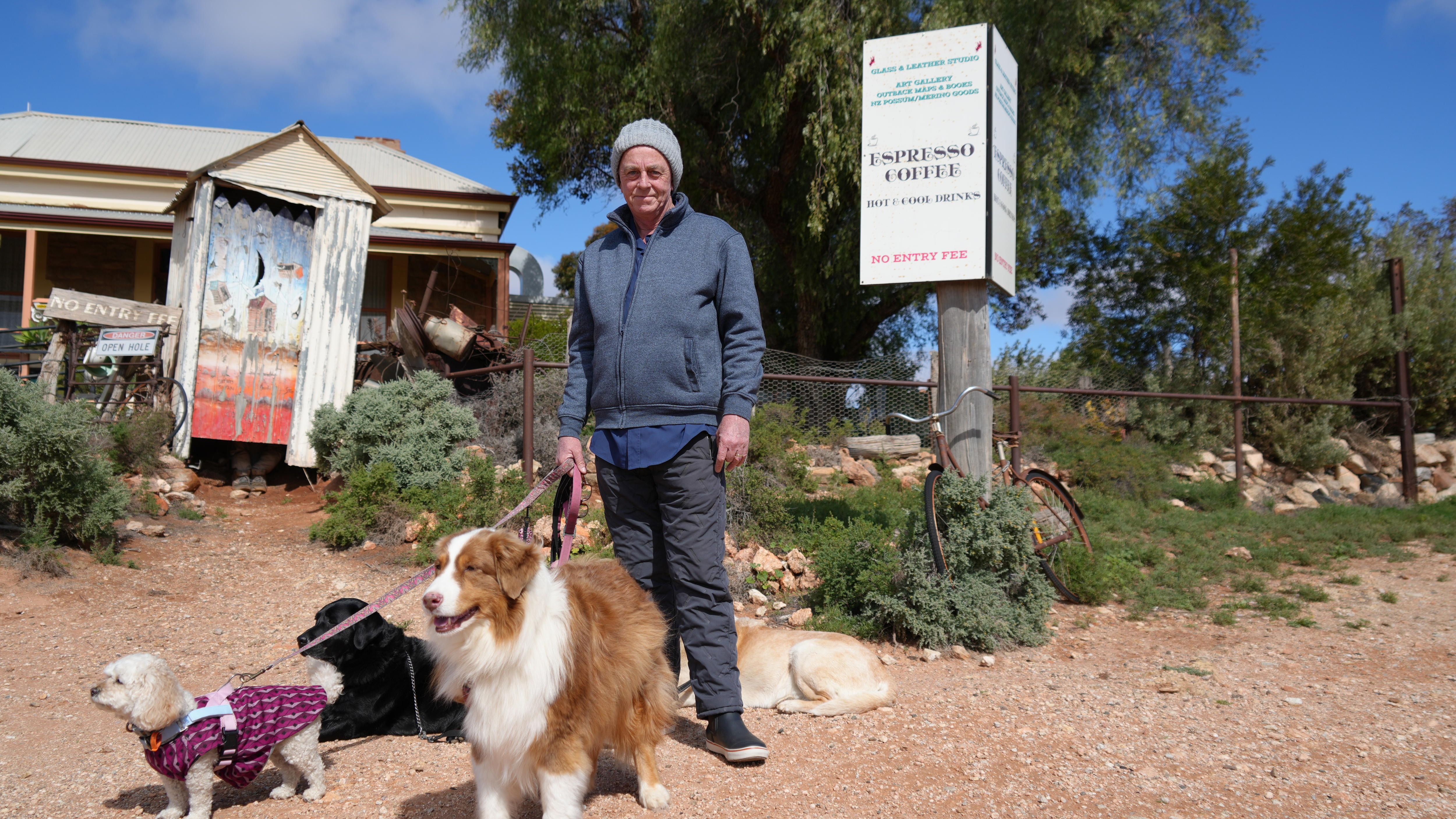 An older man wearing a beanie and jumper holding a lead attached to three dogs standing in front of a building on a dirt road.