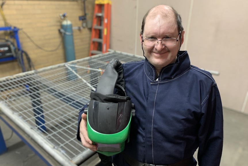 A man stands with a welding helmet, looking at the camera.