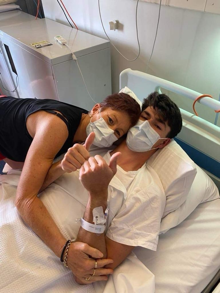 A man in a hospital bed wearing a mask holding both thumbs up being hugged by his mum.