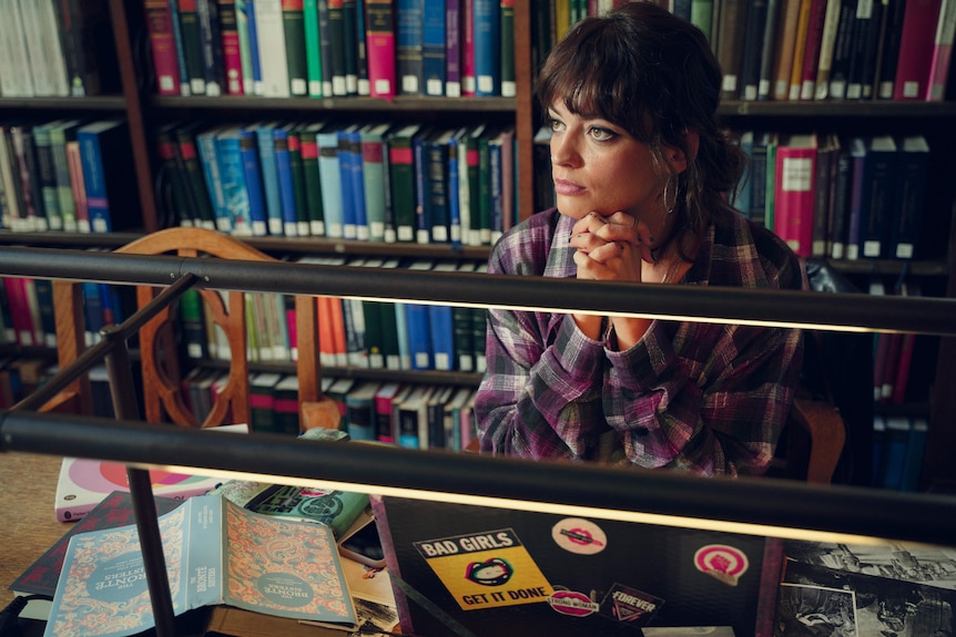 A teenage girl sits with her chin resting in her hands at the school library surrounded by books.