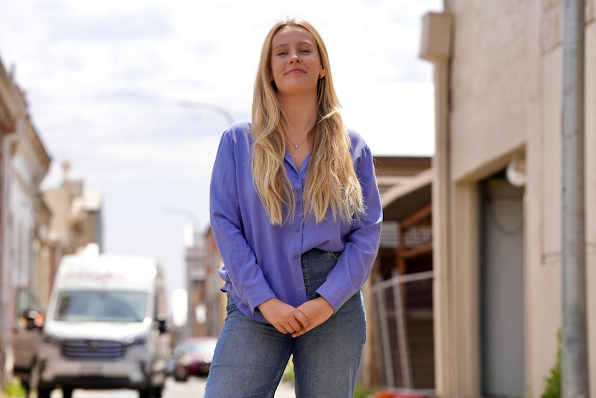 Young woman with long blonde hair and bright purple shirt half-tucked into jeans stands on street smiling.
