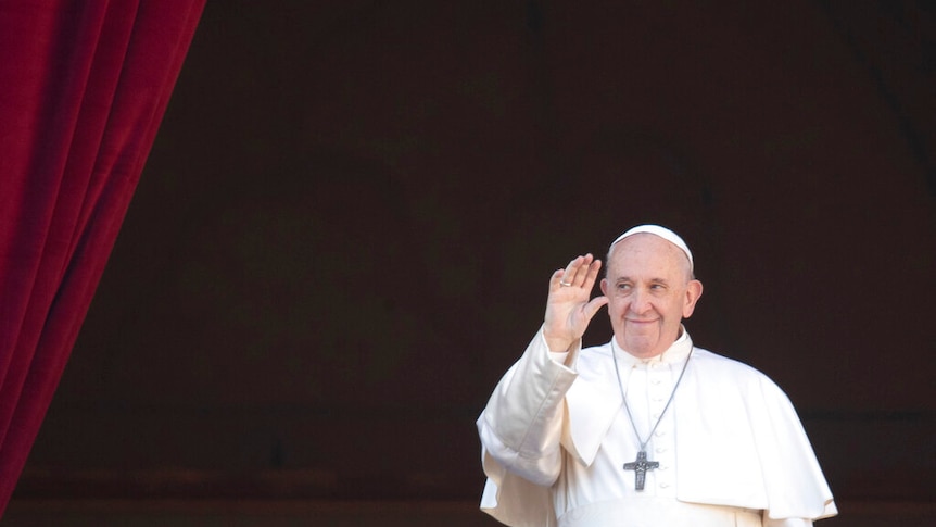 Pope Francis is pictured in white robes and waves in front of billowing red velvet curtains.