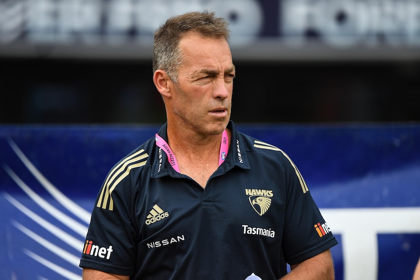 An AFL coach dressed in Hawthorn colours looks away from the camera during a game.