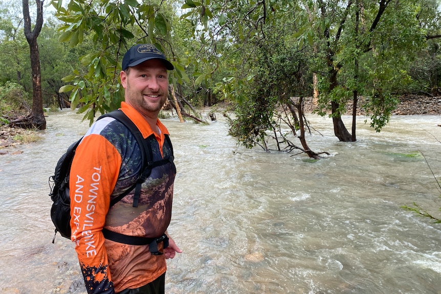 A man standing with a cap in an orange shirt in front of a creek