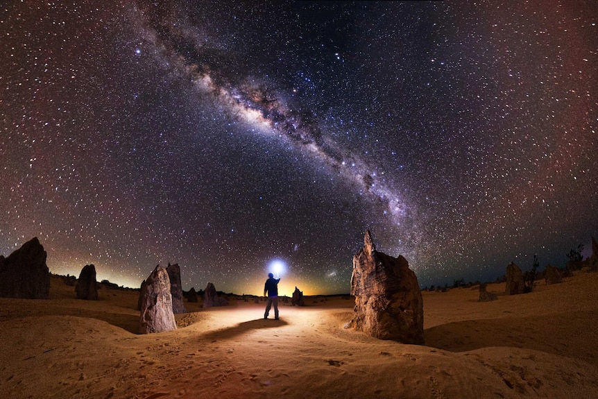 A photographer looks up to the sky beneath the stars of the Milky Way while holding a flashing device in the desert