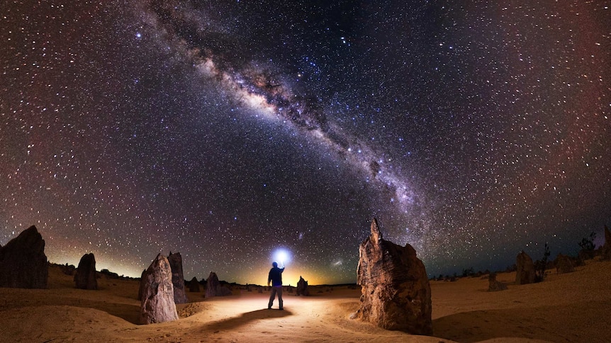 A photographer looks up to the sky beneath the stars of the Milky Way while holding a flashing device in the desert