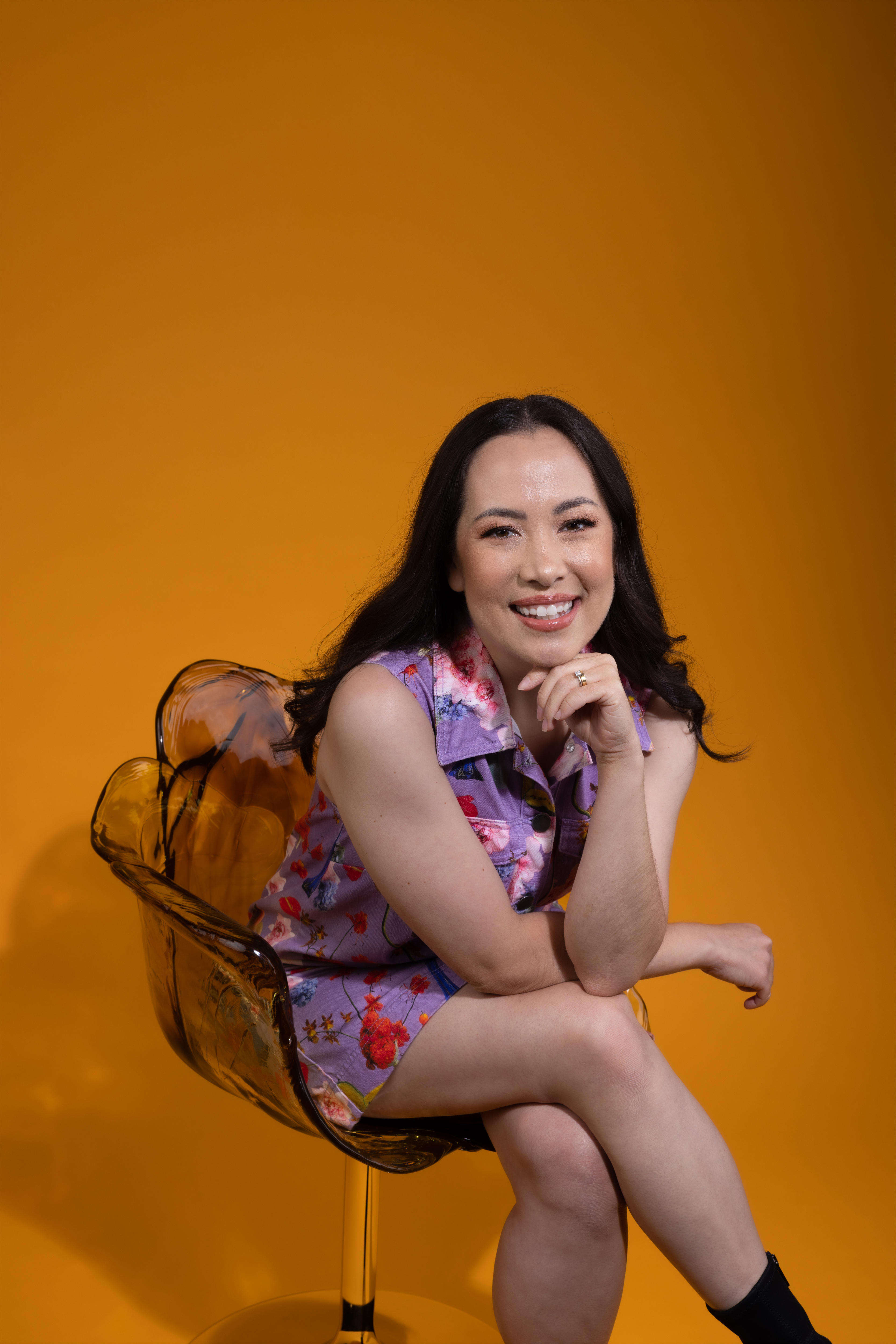 Asian Australian woman with dark hair wears purple dress with colourful splodges while seated in front of a yellow backdrop.