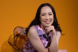 Asian Australian woman with dark hair wears purple dress with colourful splodges while seated in front of a yellow backdrop.