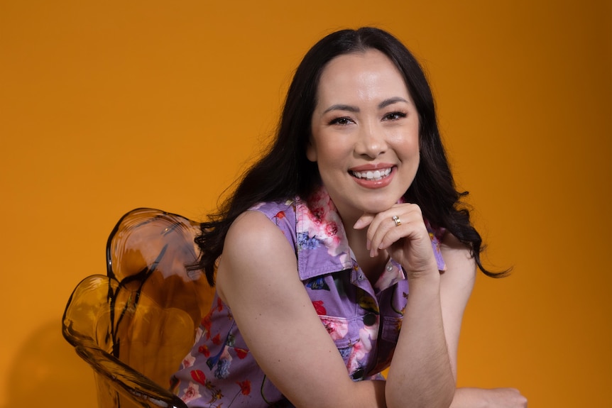 Asian Australian woman with dark hair wears purple dress with colourful splodges while seated in front of a yellow backdrop.
