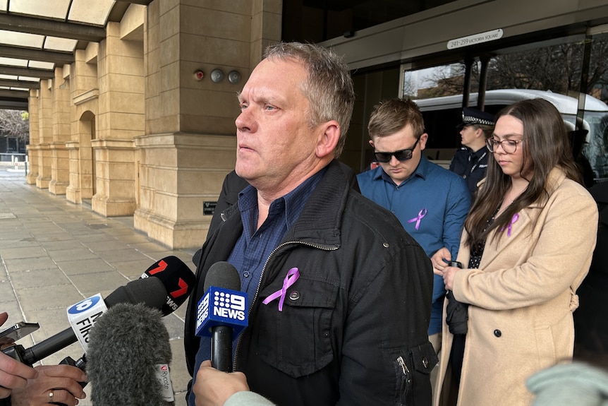 A man wearing a purple ribbon on his jacket stands with media microphones in front of him. A young man and woman are behind him