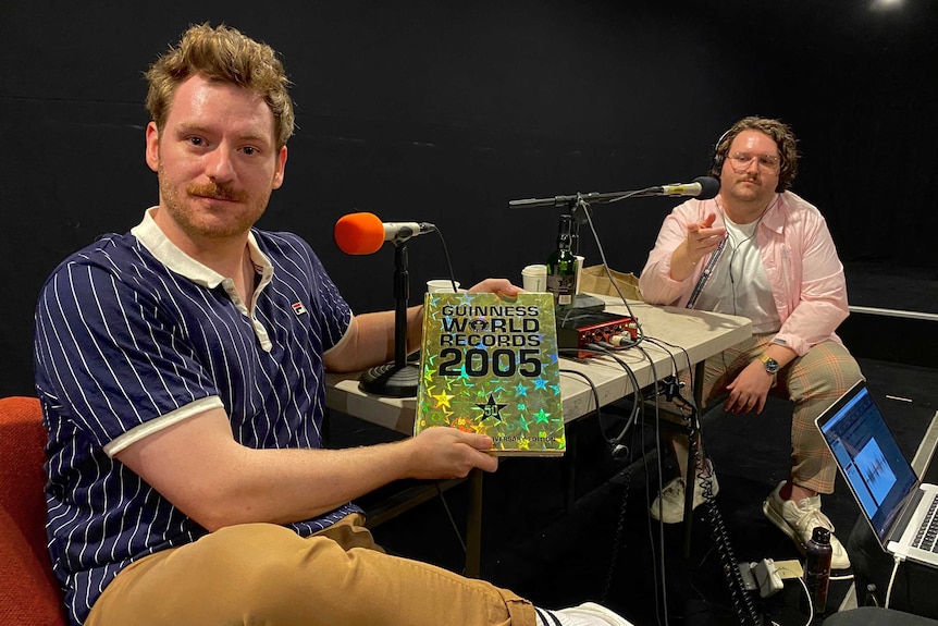 Two people sitting in front of microphones, one of them in a blue shirt holding a copy of the Guinness World Records 2005.