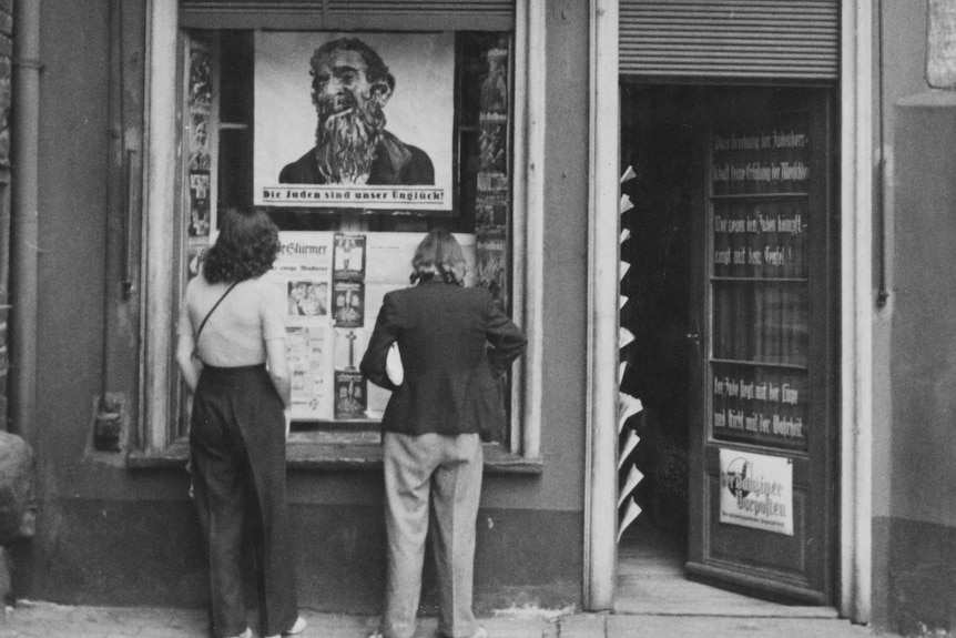 Two people stand outside a newspaper office. An illustration of a bearded jewish man is on a poster in the window.