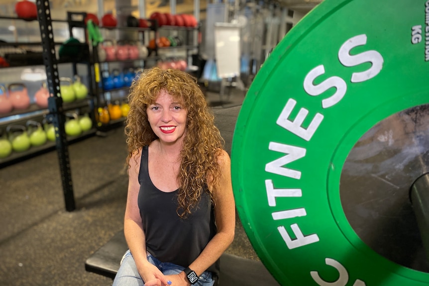 A curly-haired woman wearing a blank singlet sits in a gym, surrounded by weights.