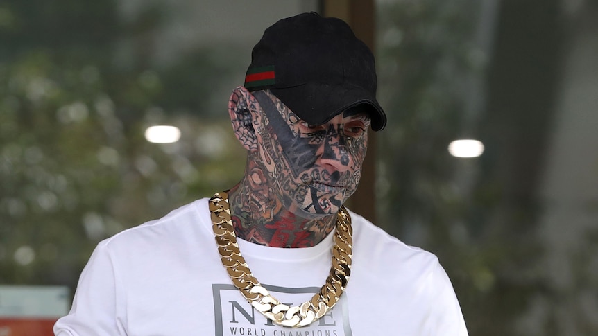A man with a face, neck and ears covered by tattoos, wearing a thick gold chain and a cap. He is looking down and to the side.