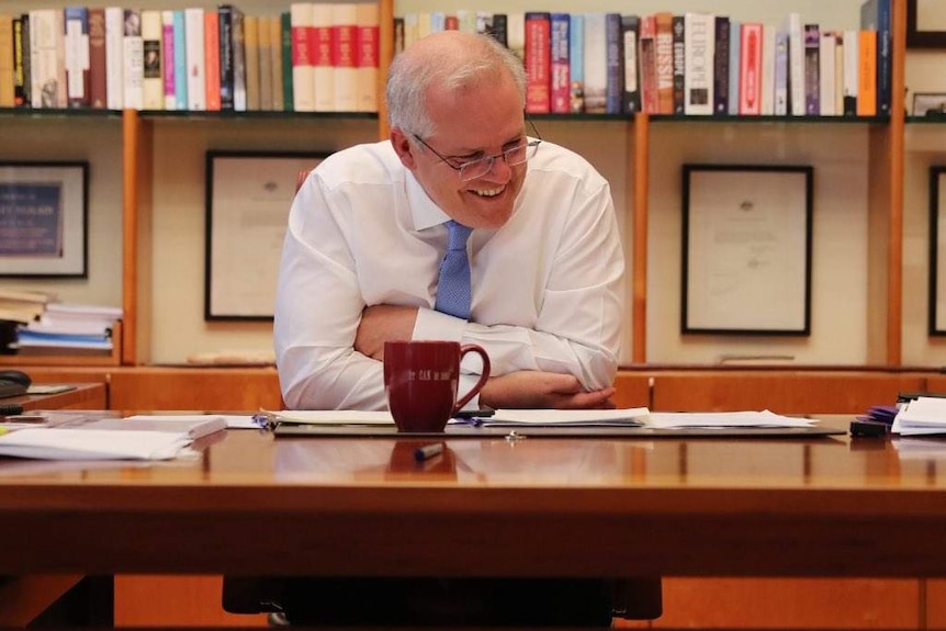 Scott Morrison sitting behind his desk with a smile on his face.