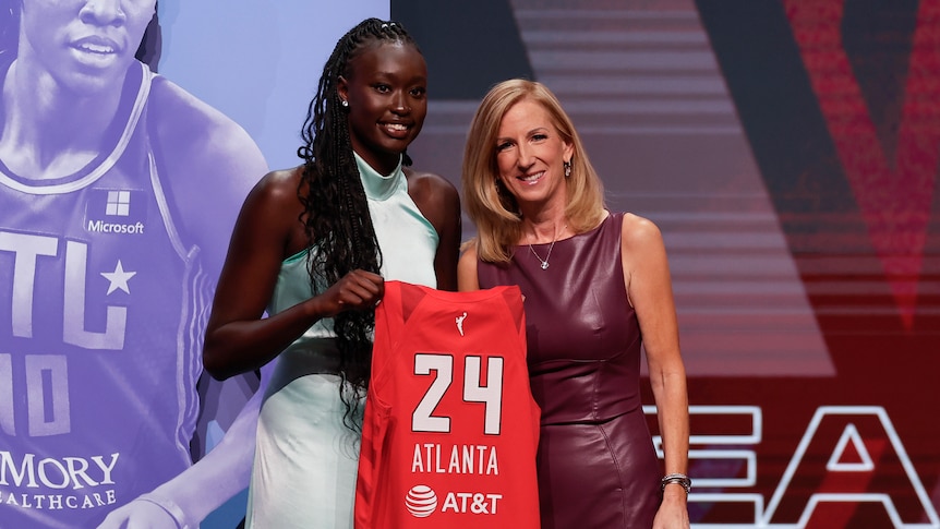 Nyadiew Puoch holds a basketball jersey with "24 Atlanta" on it after being drafted. WNBA commissioner Cathy Engelbert is there.