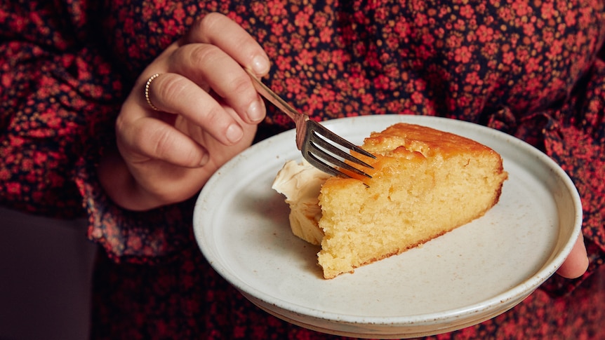 Close view of a pair of woman's hands, holding a plate with a slice of cake in one hand, sliding a fork into it with the other.