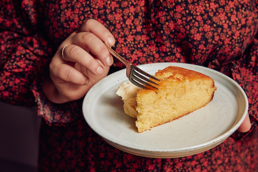Close view of a pair of woman's hands, holding a plate with a slice of cake in one hand, sliding a fork into it with the other.