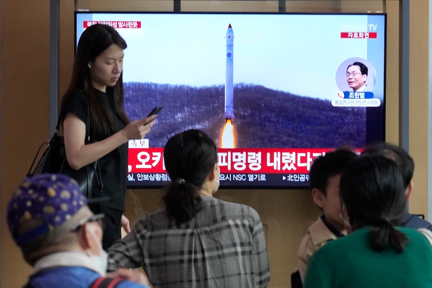 A TV screen shows a file image of a North Korea rocket launch as people watch.