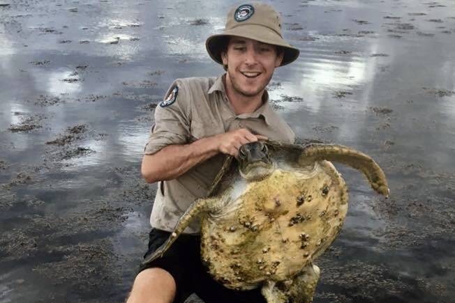 Young man wearing QPWS khaki uniform, bucket hat, holding a turtle, body of water behind.