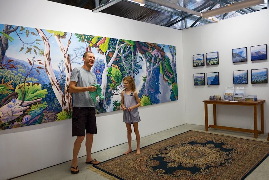 Man stands with daughter in front of a large landscape painting of a rainforest