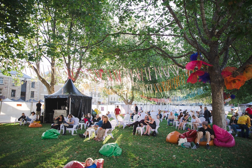 Children, men, women lounging on bean bags on green grass and trees festooned with buntings.