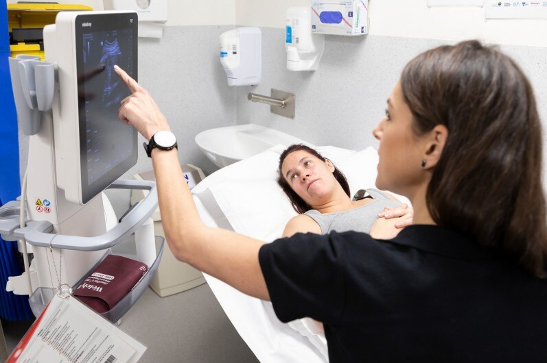 A midwife points to a scan on a small screen, as a pregnant woman on a hospital bed looks up