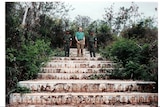 Mick Quinn stands between two Cambodian soldiers on stairs in the forest.