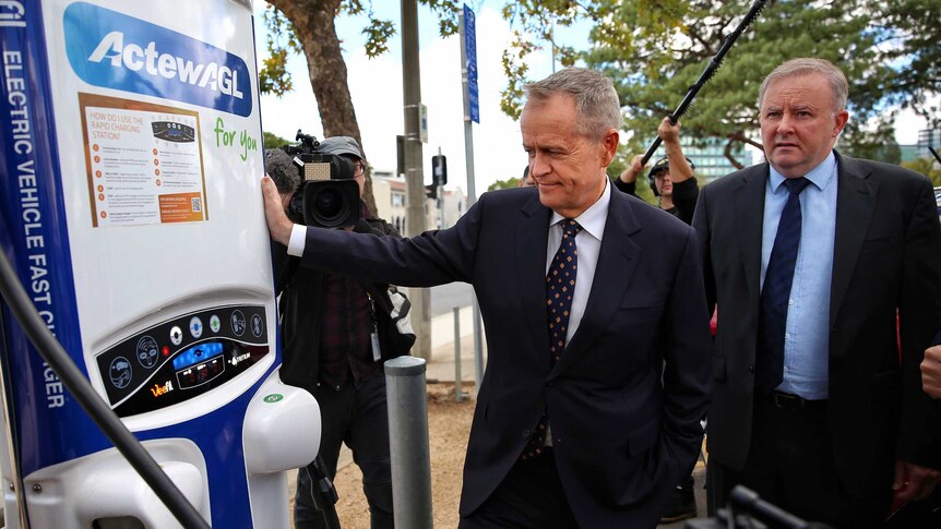 Bill shorten leans against a car recharge station as Anthony Albanese looks on