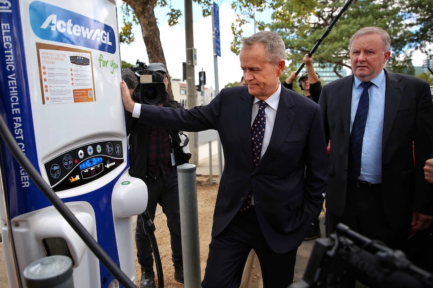 Bill shorten leans against a car recharge station as Anthony Albanese looks on