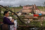 A woman holds a small white dog. A destroyed house is behind her. 