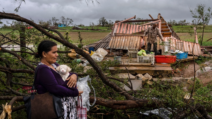 A woman holds a small white dog. A destroyed house is behind her. 