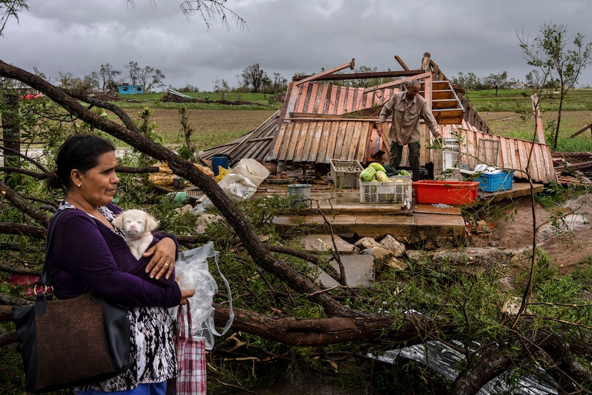 A woman holds a small white dog. A destroyed house is behind her. 