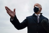 A man in a coat and face masks gestures with his right hand as he speaks to a pack of media folk.