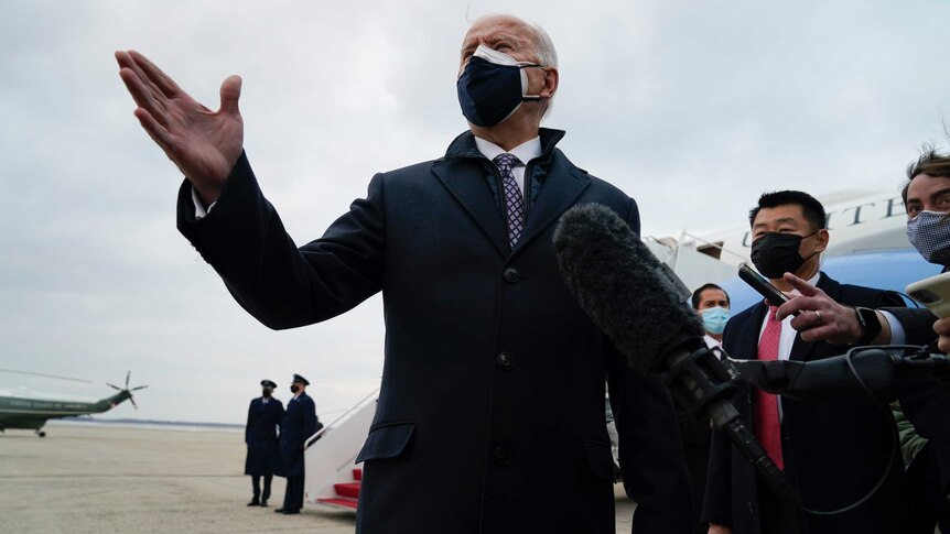 A man in a coat and face masks gestures with his right hand as he speaks to a pack of media folk.
