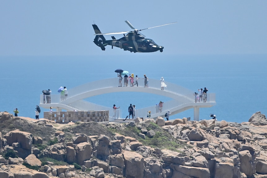 Helicopter flies past tourists on the lookout bridge next to the ocean.