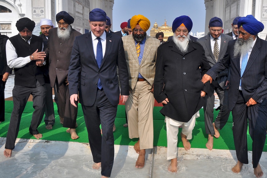 A man wearing a suit and a turban walks with a group of other men wearing suits and turbans on grass.