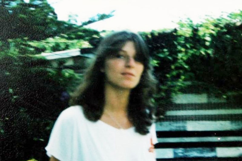 Headshot photo of Linda Reed in a white t-shirt, standing beside a garden.